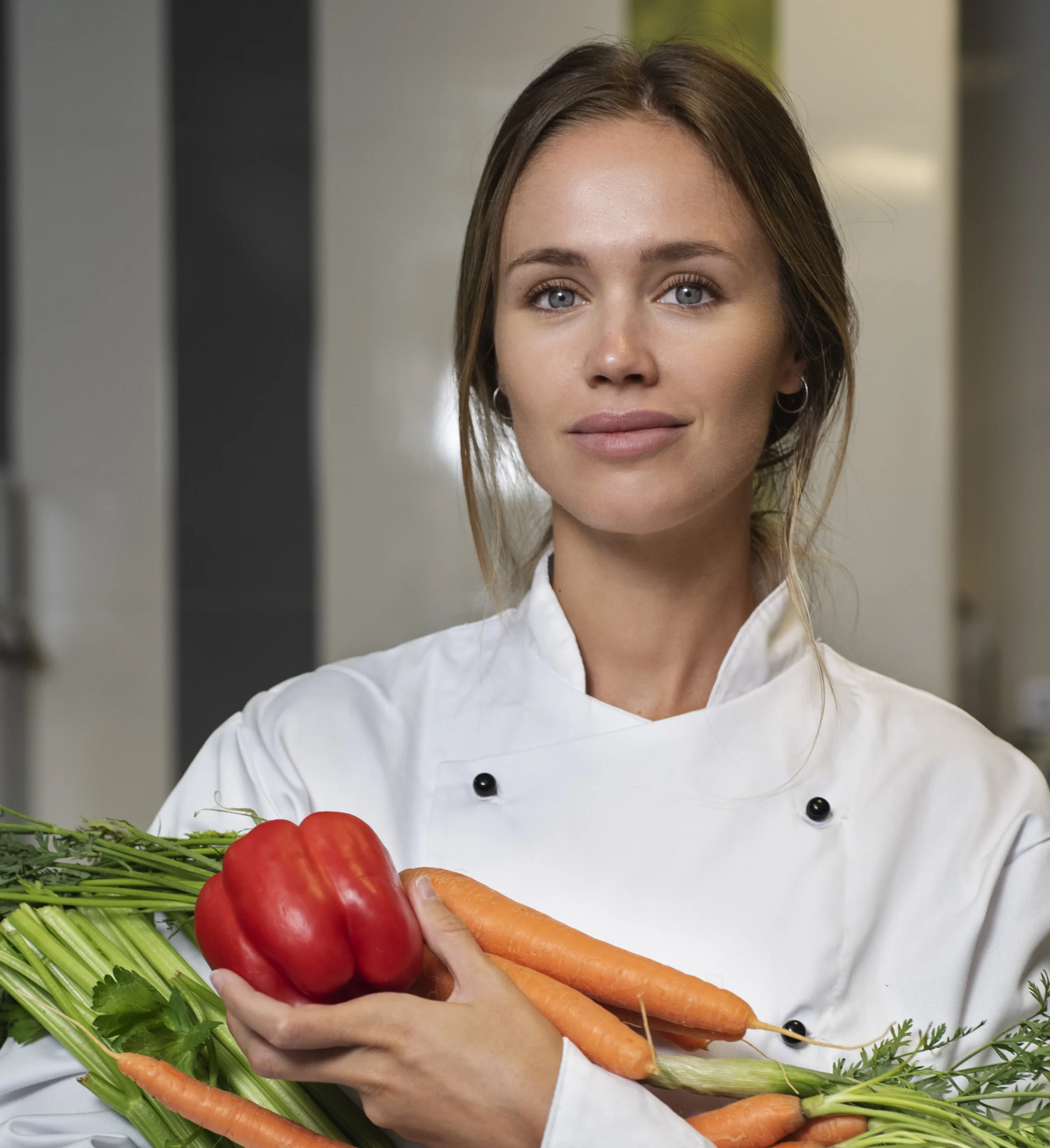 Saly abt Chef Saly standing in the kitchen wearing a white apron and holding vegetables in her arms